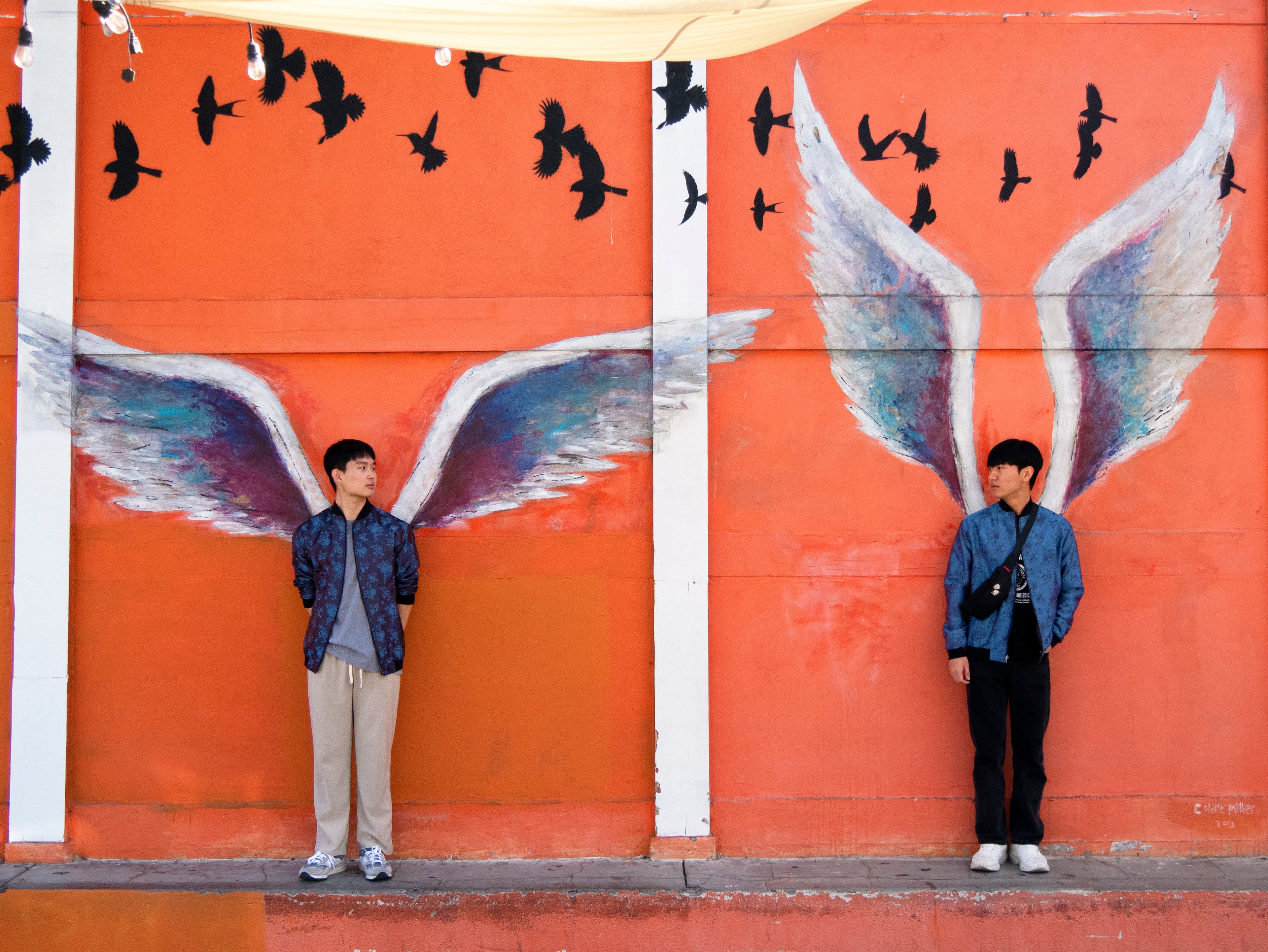 Xeon and Ryong posing in front of the angel wings in Los Angeles, each wearing a different side of the Kaelu silk bomber jacket — light grey urban style and dark grey casual look.