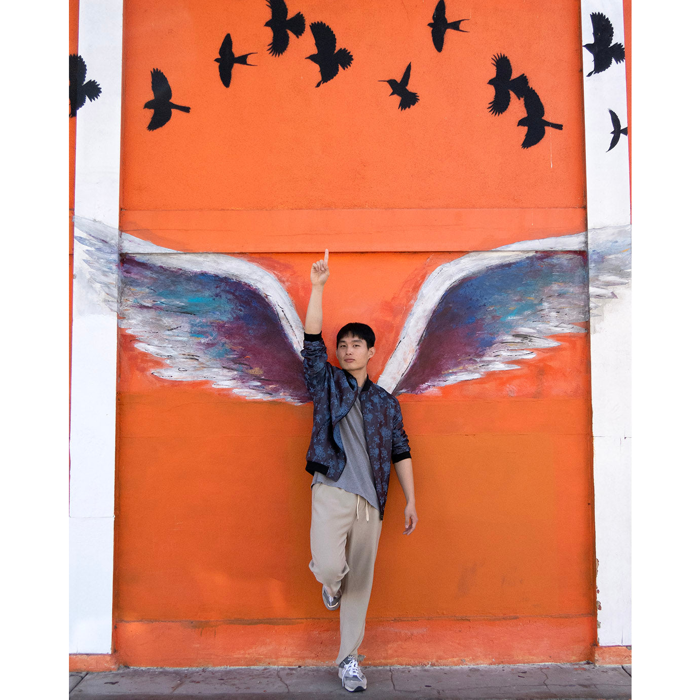 Ryong posing in front of the angel wings in Los Angeles, wearing a different side of the Kaelu silk bomber jacket — light grey urban style and dark grey casual look.
